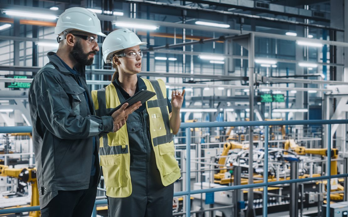 Male Specialist and Female Car Factory Engineer in High Visibility Vests Using Tablet Computer. Automotive Industrial Manufacturing Facility Working on Vehicle Production. Diversity on Assembly Plant. Male Specialist and Female Car Factory Engineer in High Visibility Vests Using Tablet Computer. Automotive Industrial Manufacturing Facility Working on Vehicle Production. Diversity on Assembly Plant.