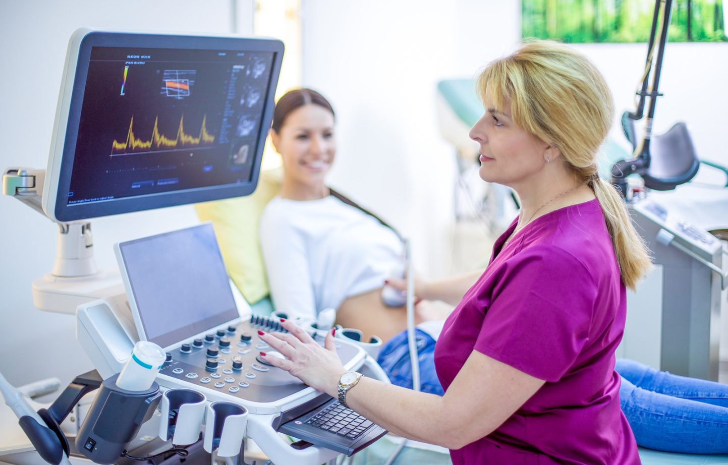 Doctor performing ultrasound on a pregnant woman. Beautiful mature female doctor in focus performing ultrasound on a pregnant smiling blurred woman in her office.